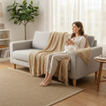 Hearthly beige knit throw blanket with geometric pattern and fringe, draped over a sofa with a woman sitting.