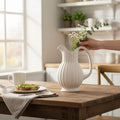 Hearthly white fluted ceramic pitcher with a subtle ribbed texture, holding wildflowers on a rustic wooden table.